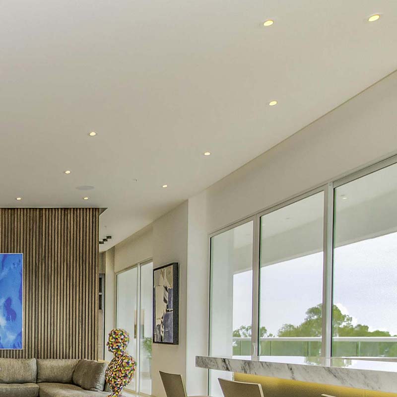 Small, round downlights installed in a white ceiling, brightening a living area with large windows and neutral décor.