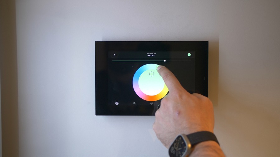 A hand touching a wall-mounted touch screen adjusting a lighting control color wheel. 