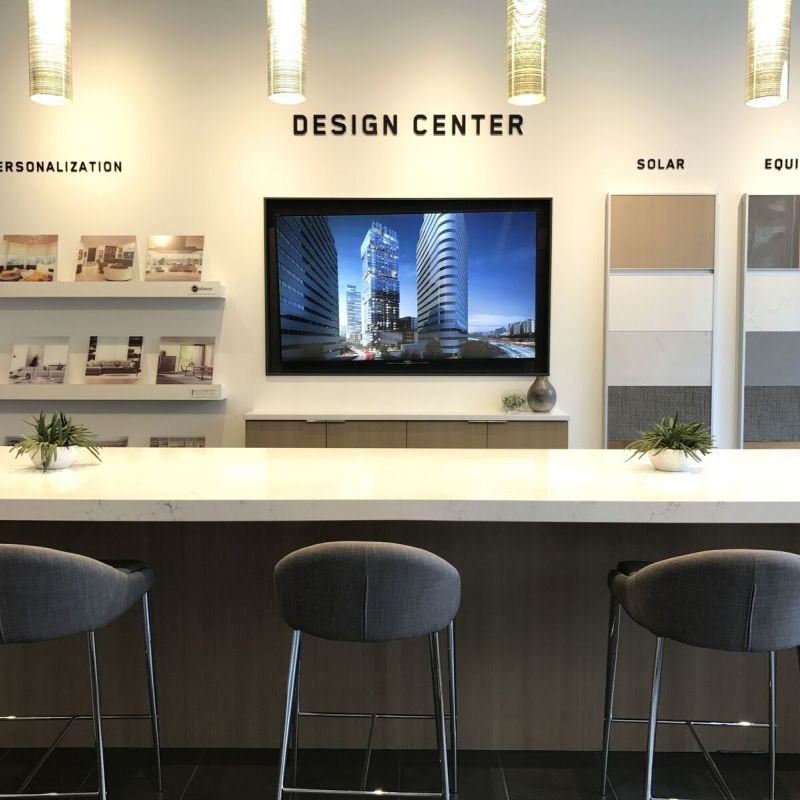 “Design Center” showroom with a long white counter and three gray barstools facing a wall-mounted screen, surrounded by sample boards, brochures, and pendant lights.